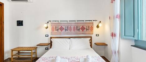 Ein Hotelzimmer mit einem Bett, zwei Nachttischen, einem Holzbeistelltisch, einer Wand-Klimaanlage und einem gemusterten Wandteppich über dem Bett.