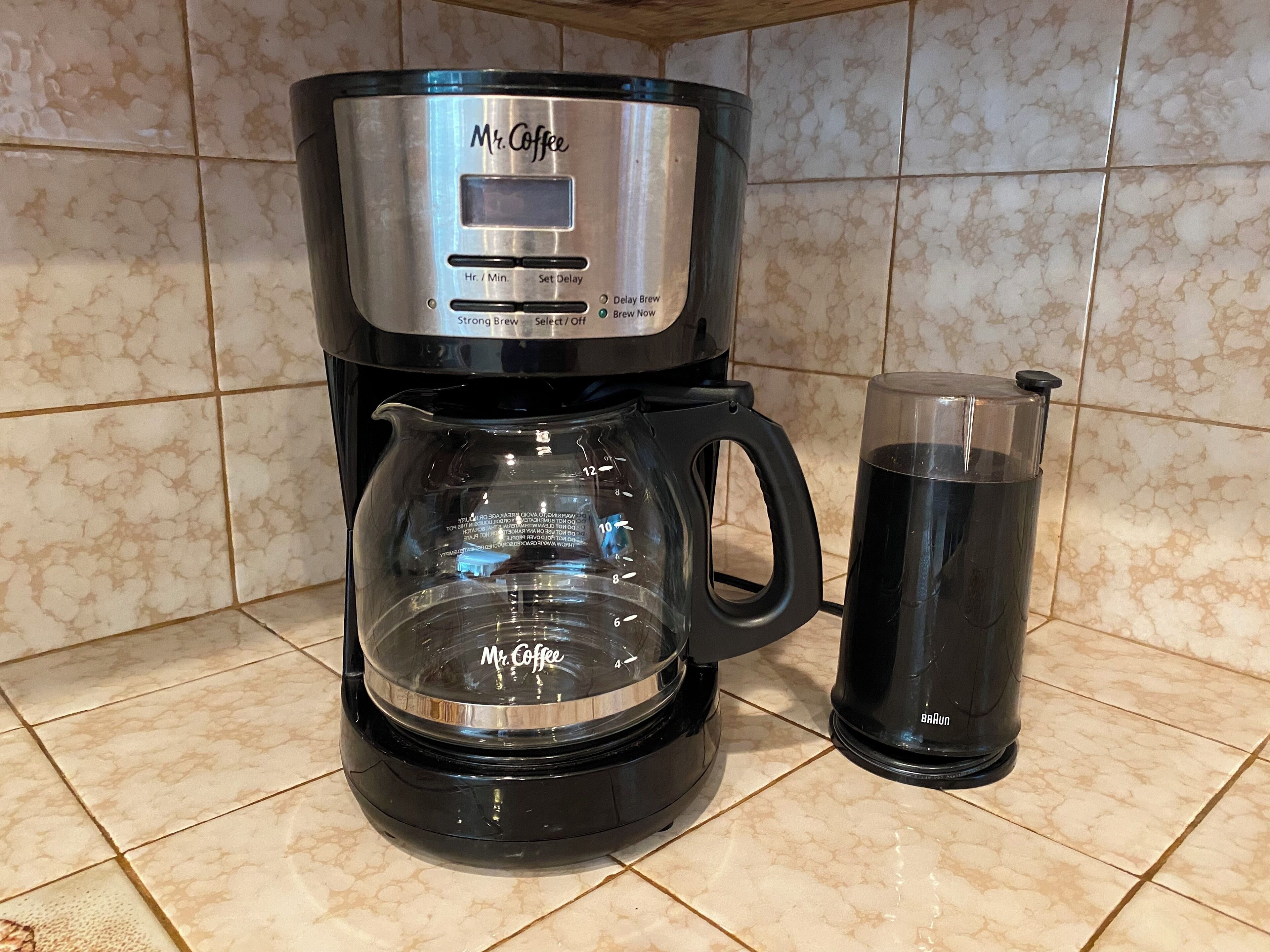 Une machine à café de la marque Mr. Coffee, dotée d’une verseuse en verre, et une cafetière Braun, posées sur un plan de travail carrelé dans la cuisine.