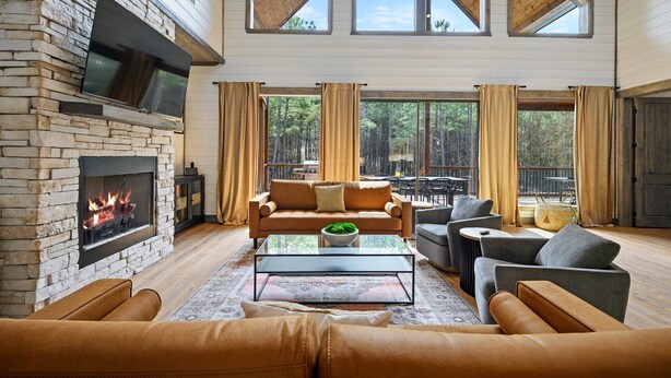 A spacious living room with a stone fireplace, leather sofas, and a large window overlooking a forest.
