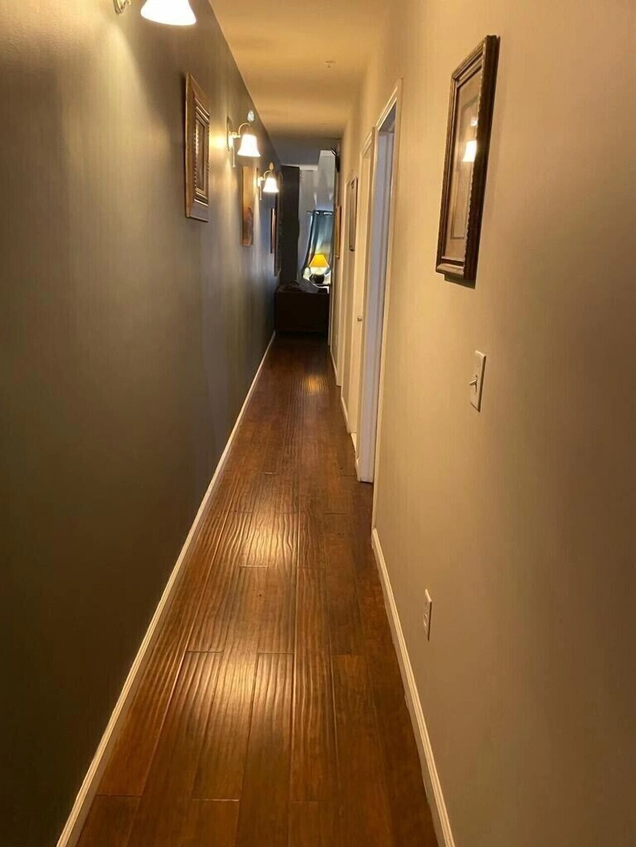 A hallway with wooden flooring, light-colored walls, and wall-mounted light fixtures.