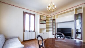 A dining area with a wooden table and chairs, a sofa, a TV, and built-in shelving. - YiD Amaryllis (Firenze)