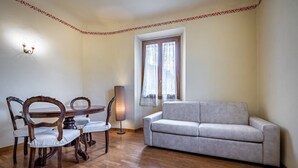 A room with a dining table and chairs, a sofa, a lamp, and a window with curtains. - YiD Amaryllis (Firenze)