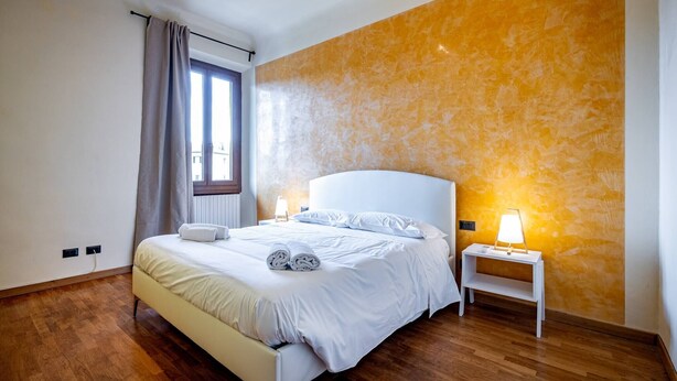 A bedroom with a bed, a nightstand, a lamp, and a window with curtains. - YiD Amaryllis (Firenze)
