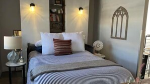 A bedroom with a bed, a bedside table with a lamp, a small table with a decorative item, and a wall-mounted shelf with books and a picture frame. - Charming 1-bedroom apartment with WiFi, AC in wonderful Denver (Denver)