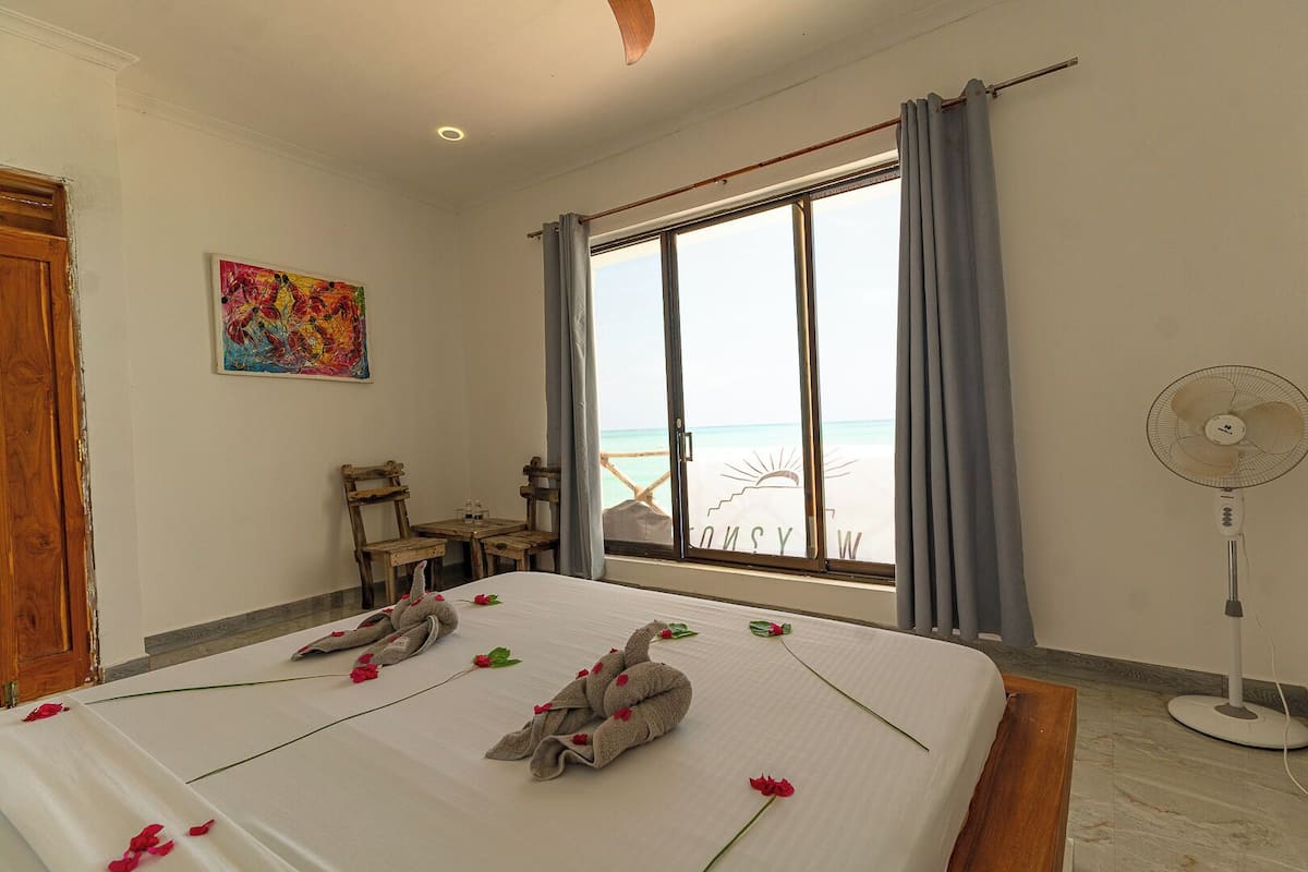 Habitación de hotel con cama, escritorio, silla, ventilador y ventana con vista a la playa.