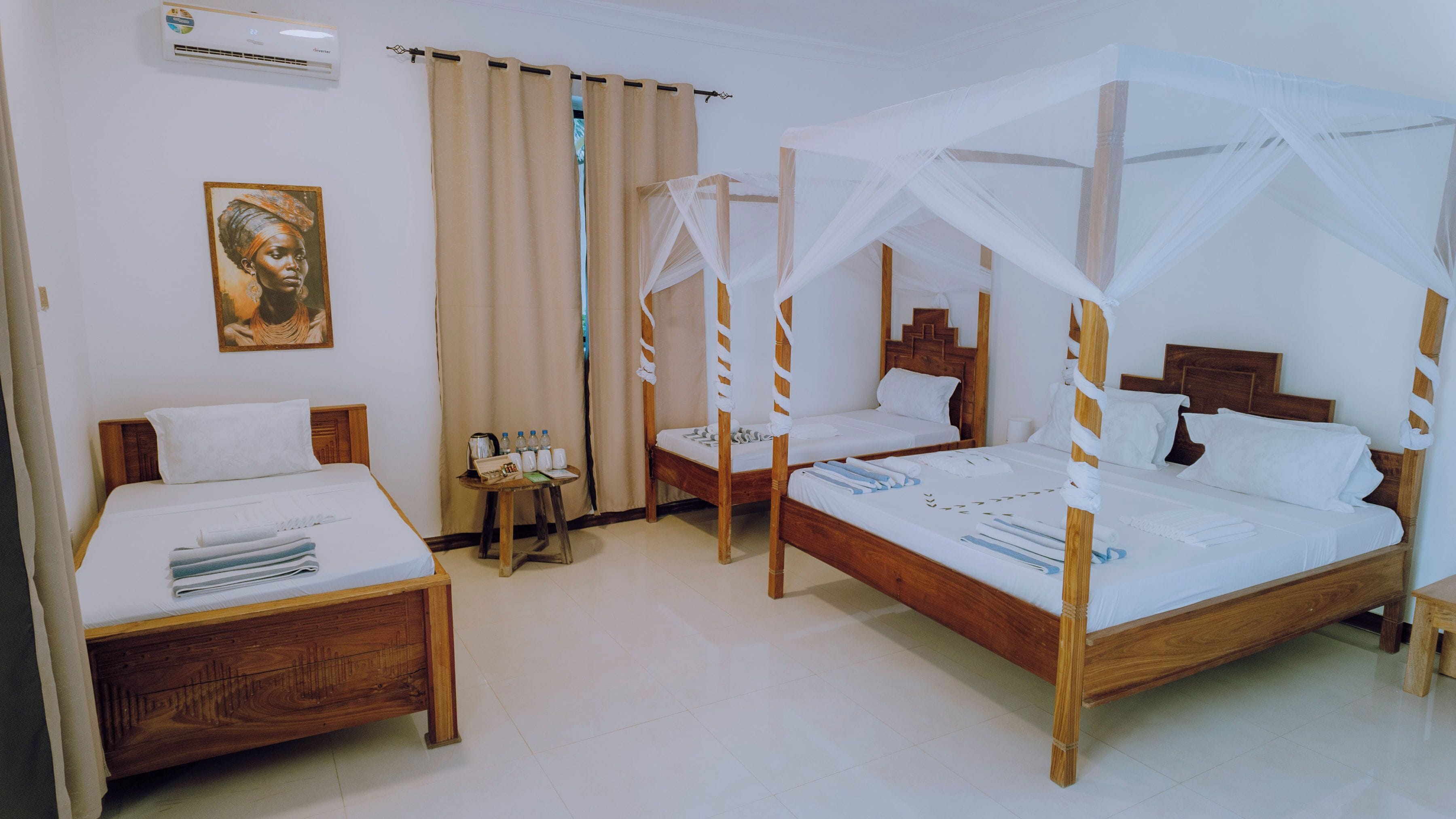Ein Hotelzimmer mit zwei Holzbett, eines mit Baldachin und eines ohne. Es gibt einen kleinen Tisch mit einem Wasserkocher und Flaschen, sowie ein Gemälde an der Wand.