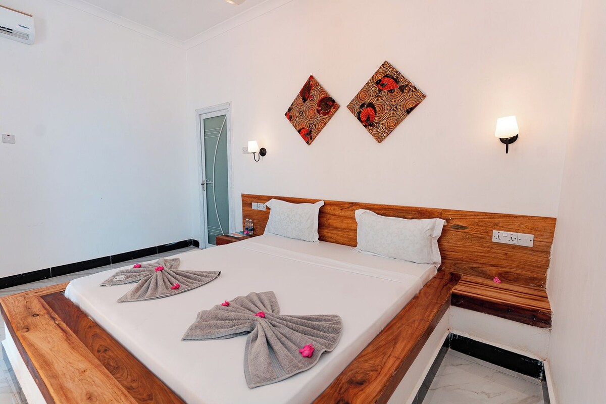 A hotel room with a wooden headboard, a bed with white linens, two wall-mounted lamps, and a door leading to a bathroom.