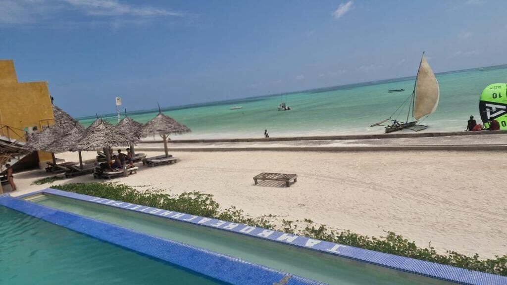 Una playa con piscina, sombrillas y un velero.