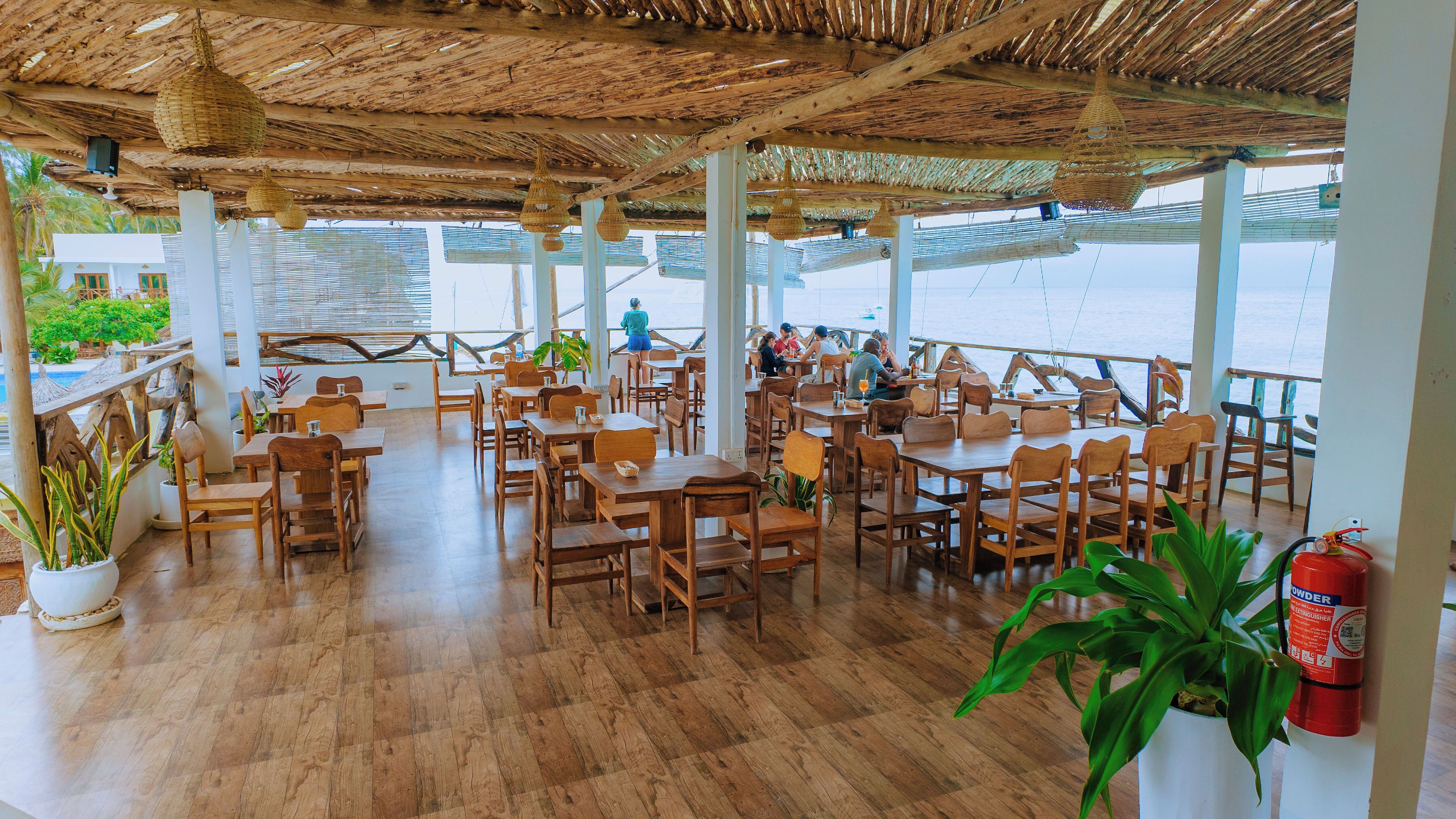 An outdoor dining area with wooden tables and chairs, a thatched roof, and a view of the sea.