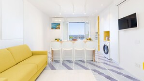 Un espace de vie compact comprenant un canapé jaune, un coin repas avec quatre chaises, un lave-linge et un téléviseur fixé au mur.