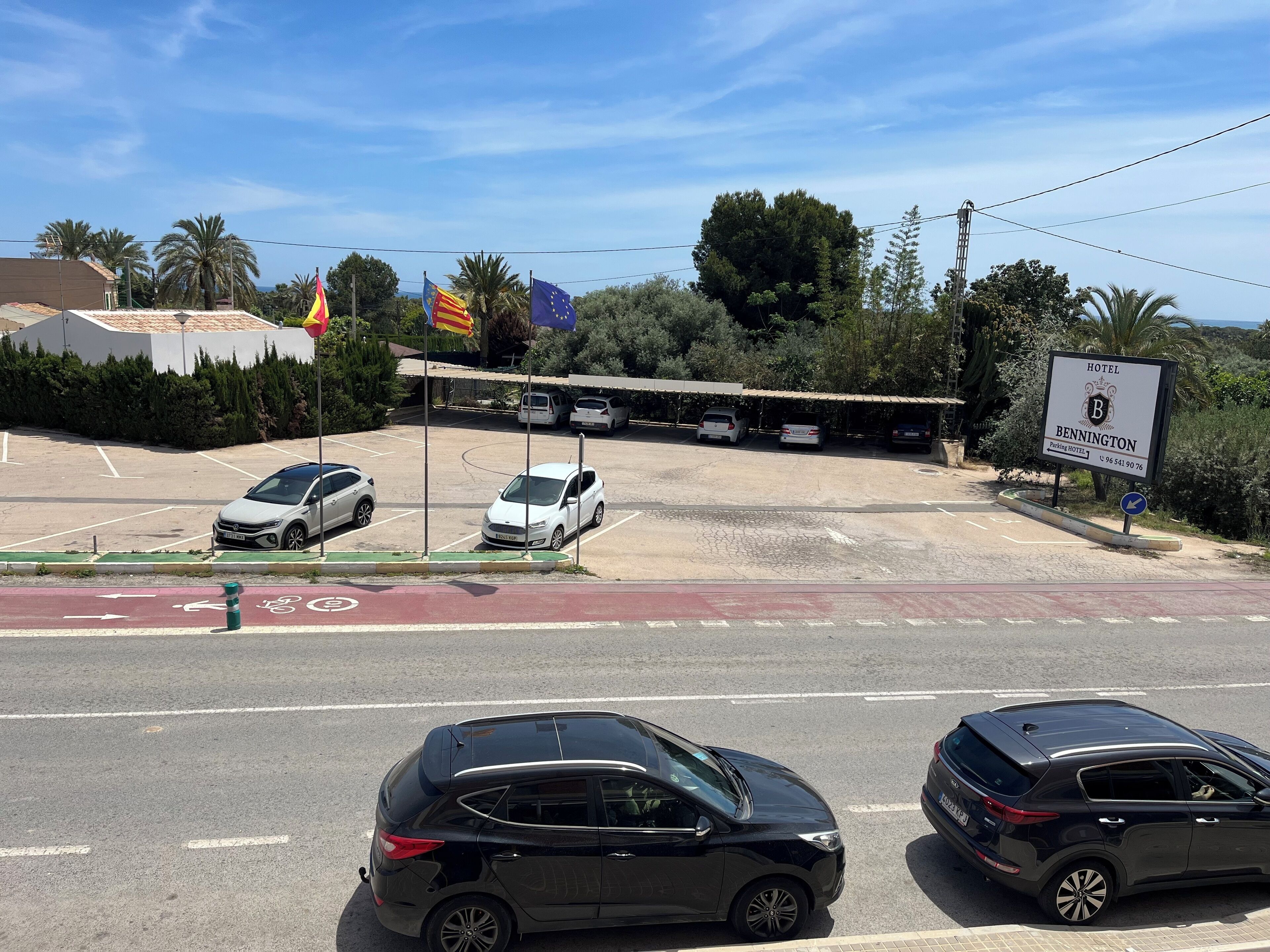 A hotel parking lot with several cars, a bike lane, and a sign indicating the hotel name and contact number.