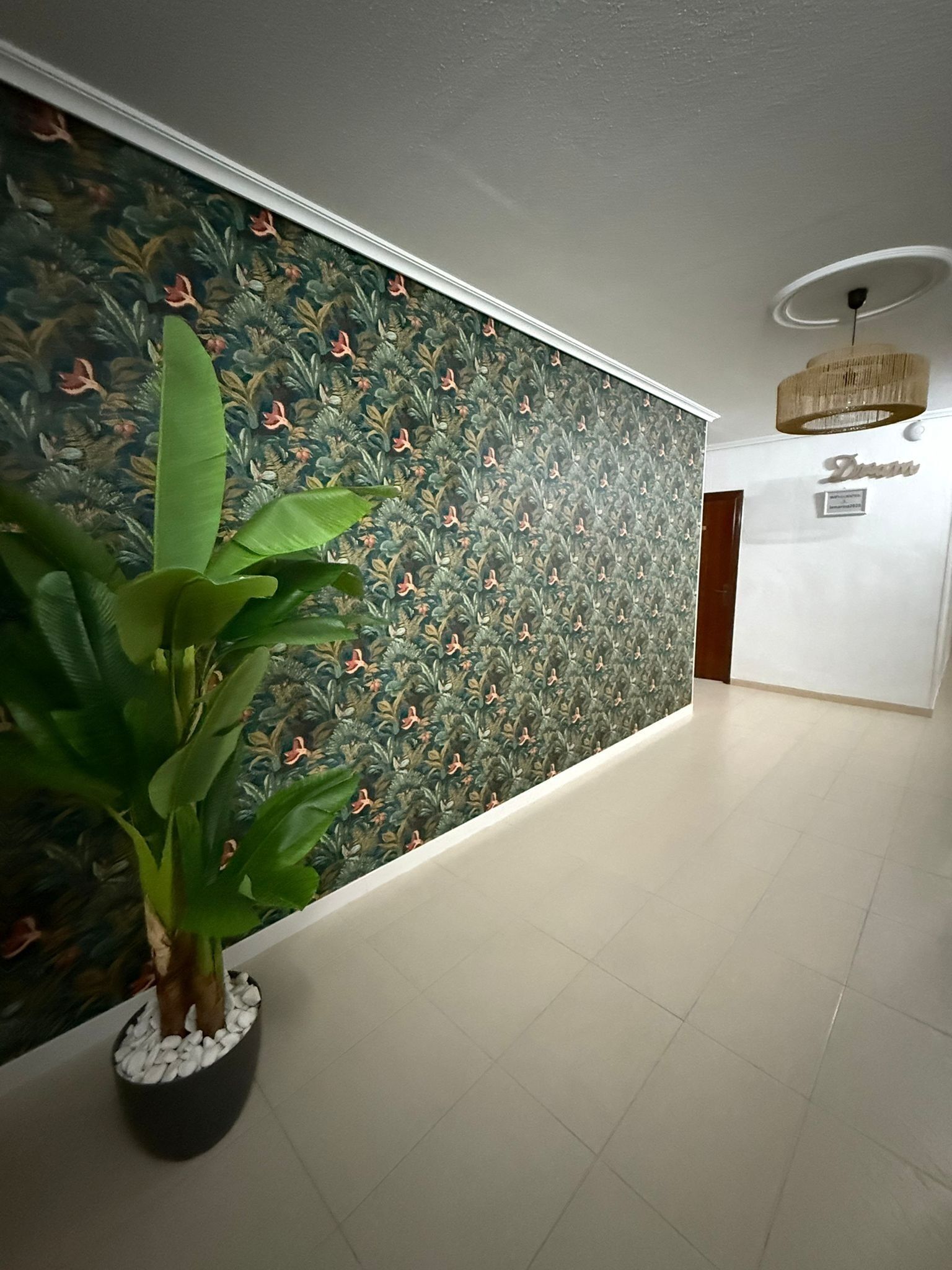 Un couloir orné d’un papier peint à motifs floraux, avec une plante en pot et un panneau indiquant « Restaurant ».