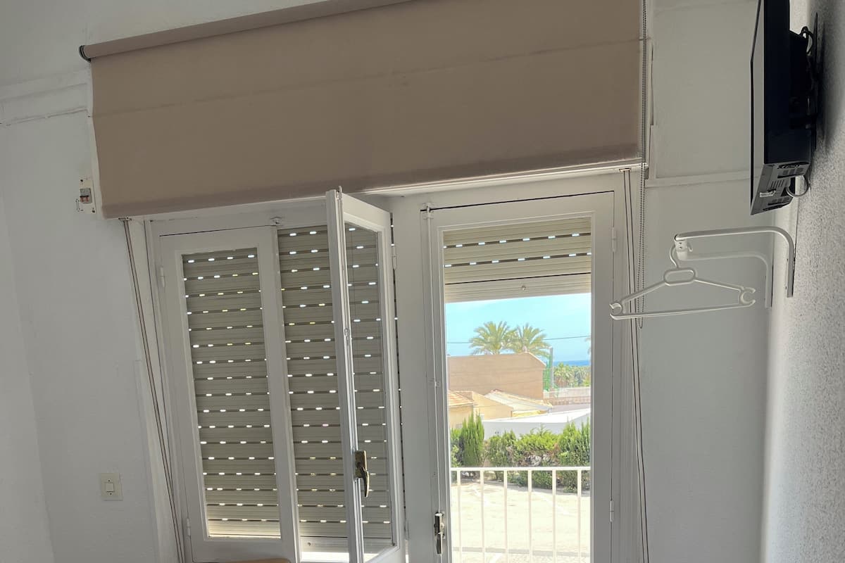A room with a bed, a chair, a television, and a door leading to a balcony with a view of palm trees and the sea.