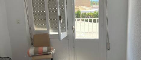 A room with a bed, a chair, a television, and a door leading to a balcony with a view of palm trees and the sea.