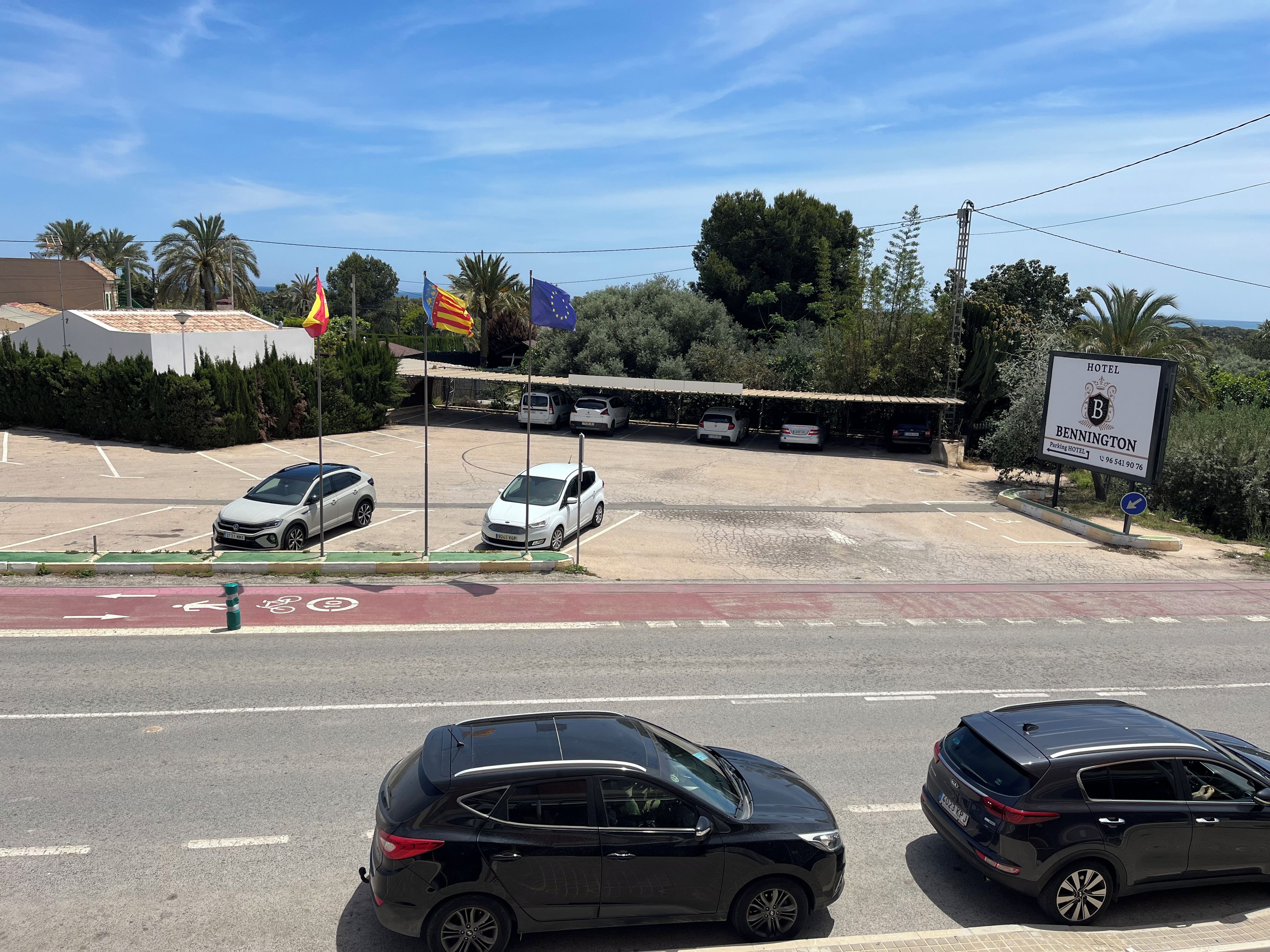 A hotel parking lot with multiple cars, a bicycle lane, and a sign indicating the hotel's name and contact number.