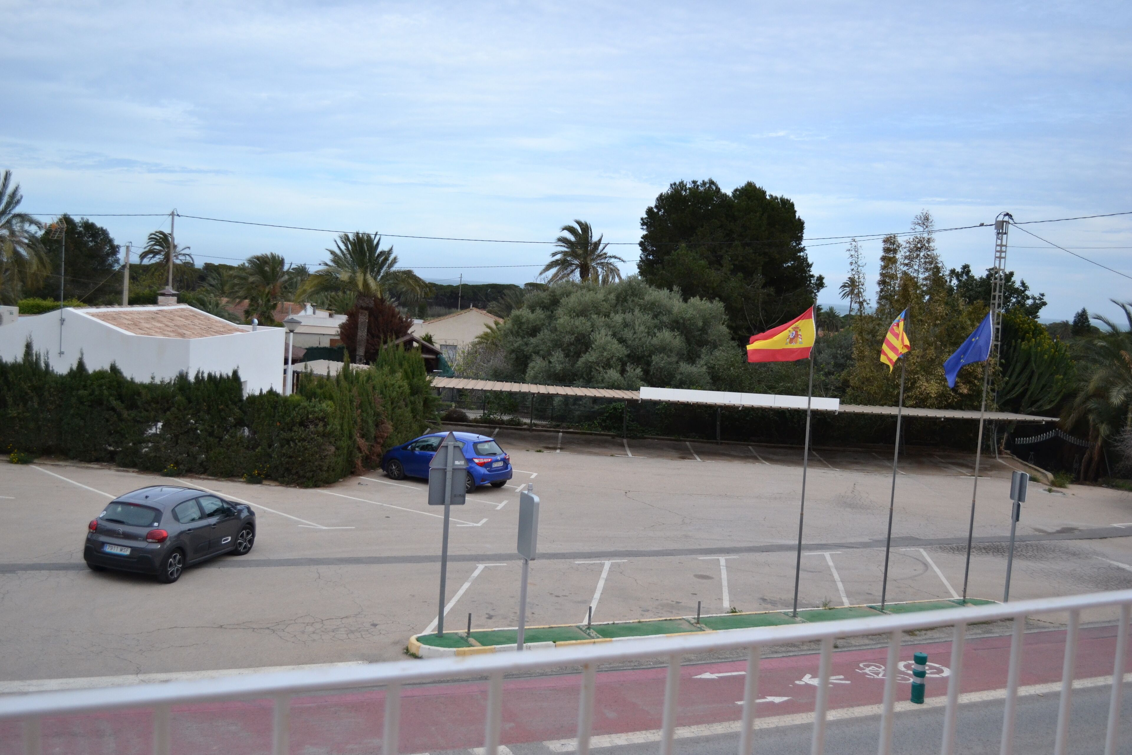 Un estacionamiento con varios coches, un carril bici verde y banderas de España y de la Unión Europea.