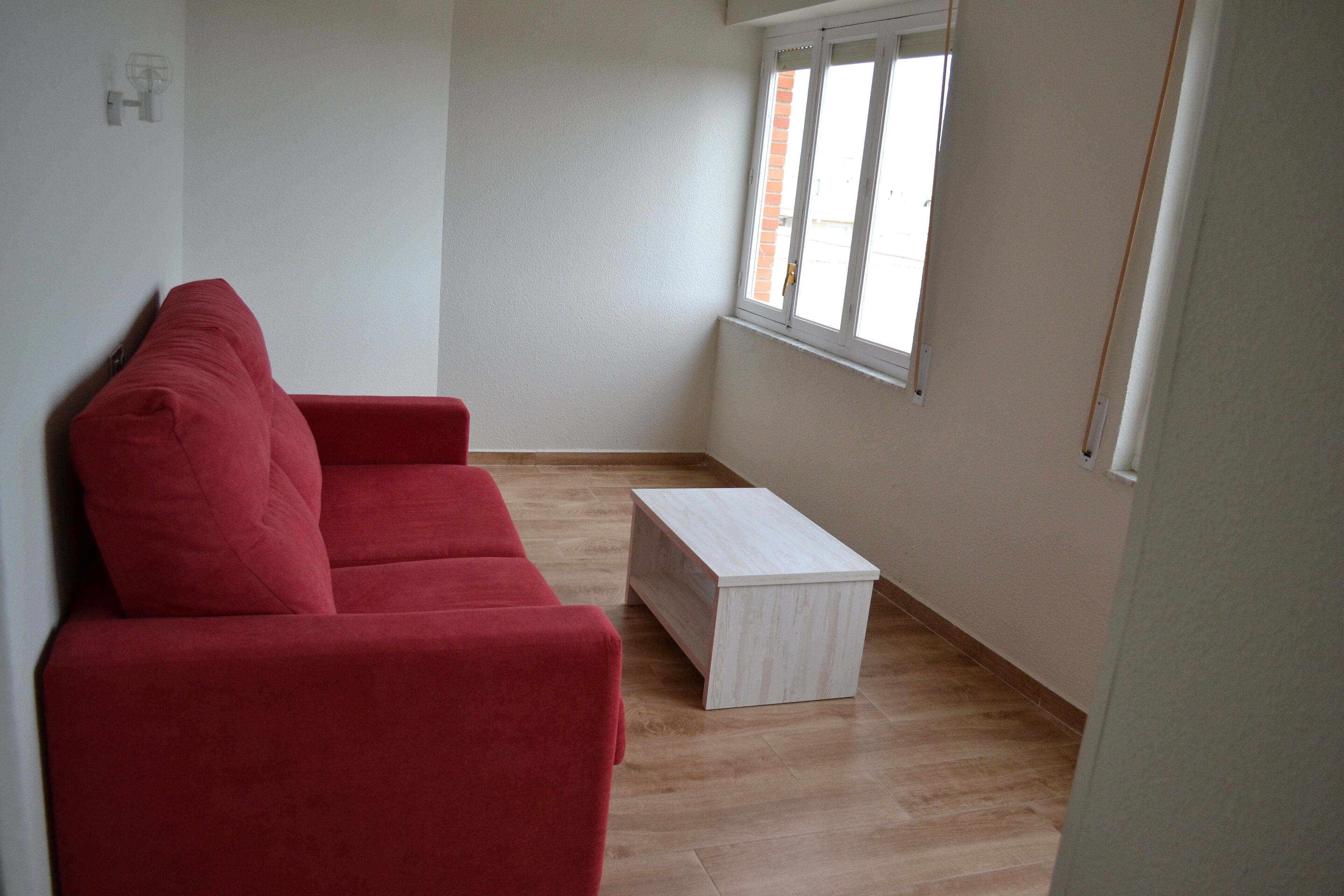 Una habitación con un sofá rojo, una mesita auxiliar blanca y una ventana con persianas.
