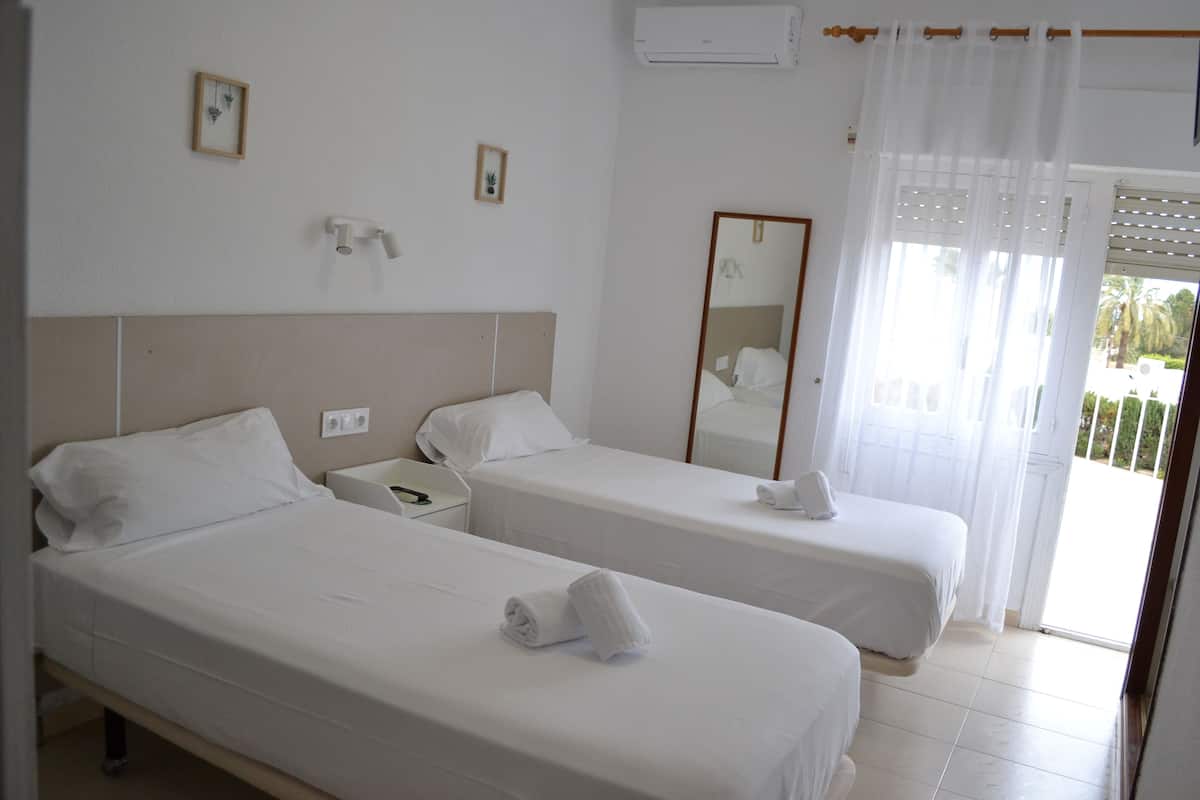 A hotel room with two single beds, a bedside table with a phone, a mirror, and a window with white curtains.