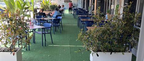 Un café al aire libre con mesas y sillas, plantas en macetas y una pared de piedra.