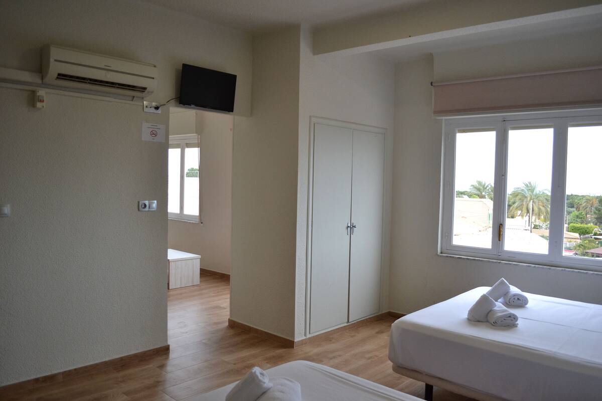 A hotel room with a bed, a TV mounted on the wall, a wardrobe, and a window with a view of palm trees.
