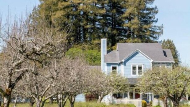 Une maison avec une clôture blanche en bois, une cheminée et un grand arbre devant.