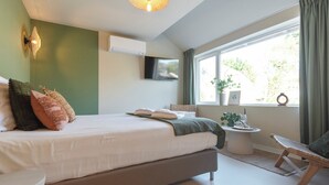 Ein modernes Schlafzimmer mit einem großen Bett, einem an der Wand montierten Fernseher, einem kleinen runden Tisch mit Pflanze und einem Fenster mit Vorhängen.