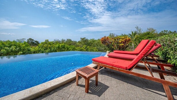Una piscina infinita de color azul claro con sillas reclinables rojas y una mesa de madera, rodeada de exuberante vegetación y vistas al océano.