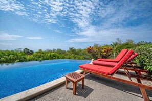 Una piscina infinita de color azul claro con sillas reclinables rojas y una mesa de madera, rodeada de exuberante vegetación y vistas al océano.