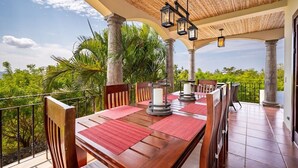 Un área de comedor al aire libre con mesa y sillas de madera, rodeada de abundante vegetación y una vista despejada del cielo.