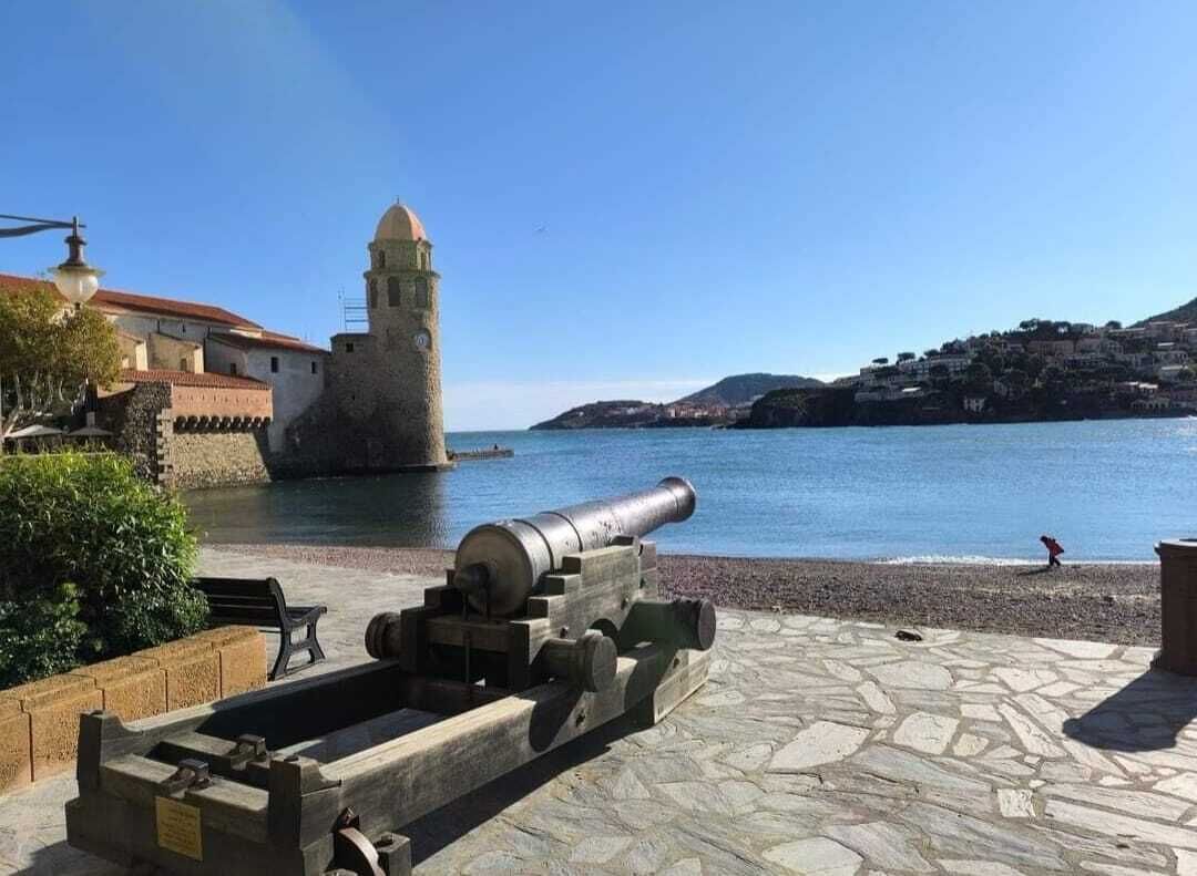 Un cañón histórico sobre una plataforma de piedra junto a la orilla, con un edificio y una torre al fondo.