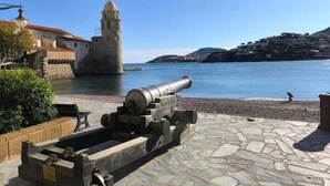 A historic cannon on a stone platform by a waterfront, with a building and a tower in the background.
