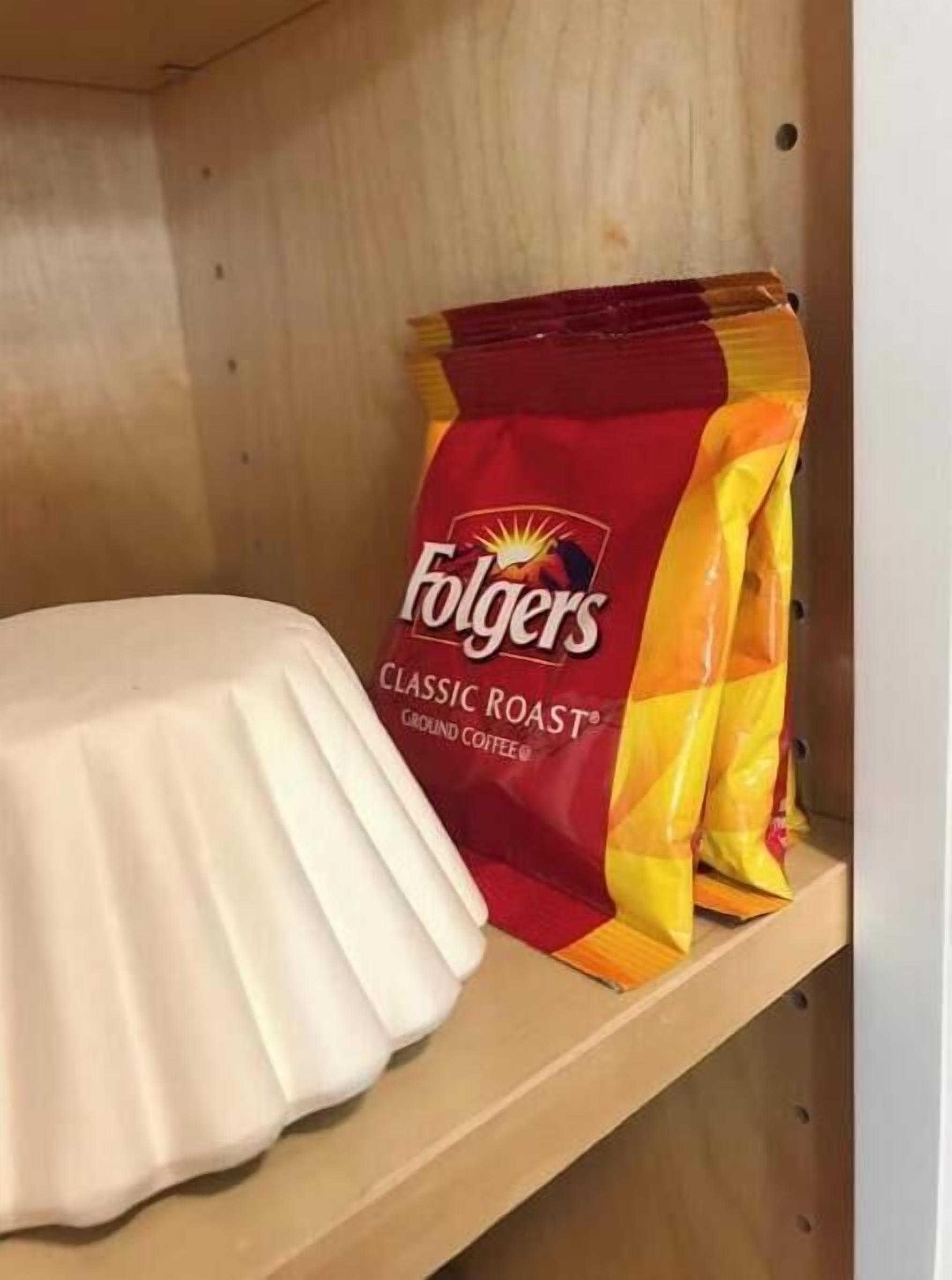 Een houten plank met een witte keramieken pot en een zak Folgers Classic Roast gemalen koffie.