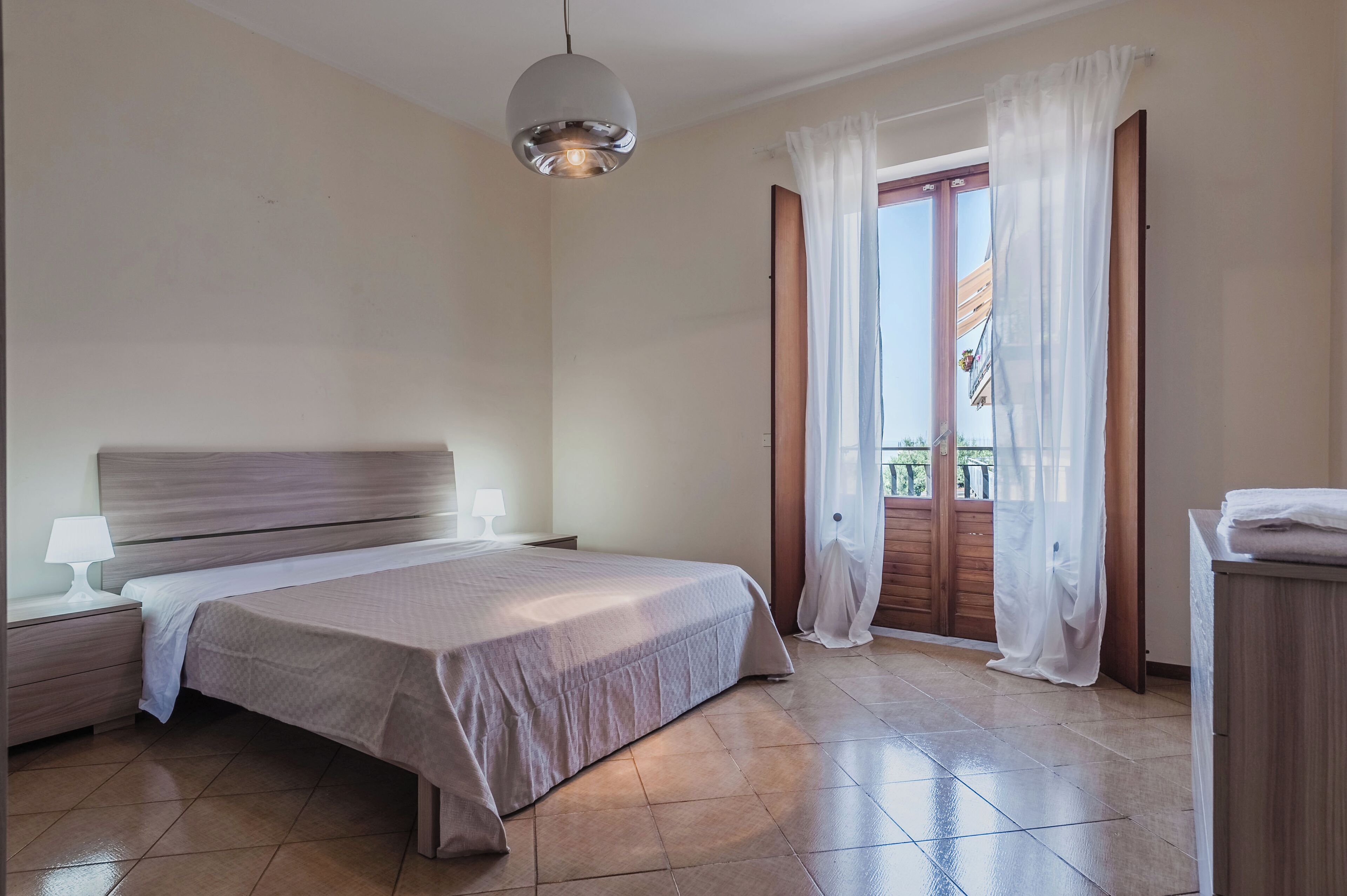 Una camera da letto con un letto, comodini, una lampada, una porta in legno e vista attraverso le tende.