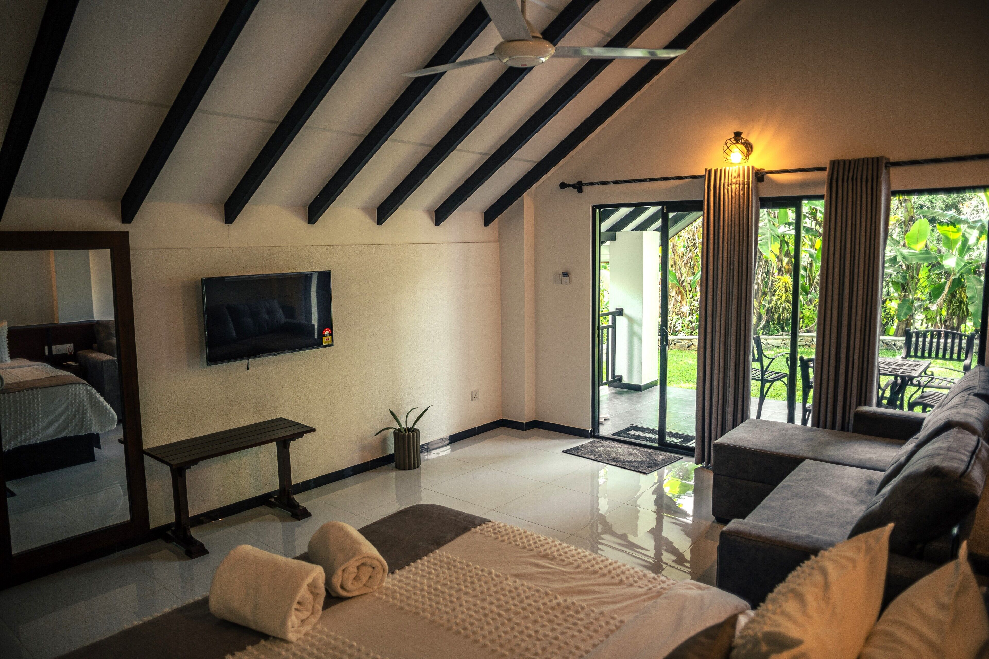 A modern hotel room with a large bed, a sofa, a TV, and a balcony with a view of greenery.