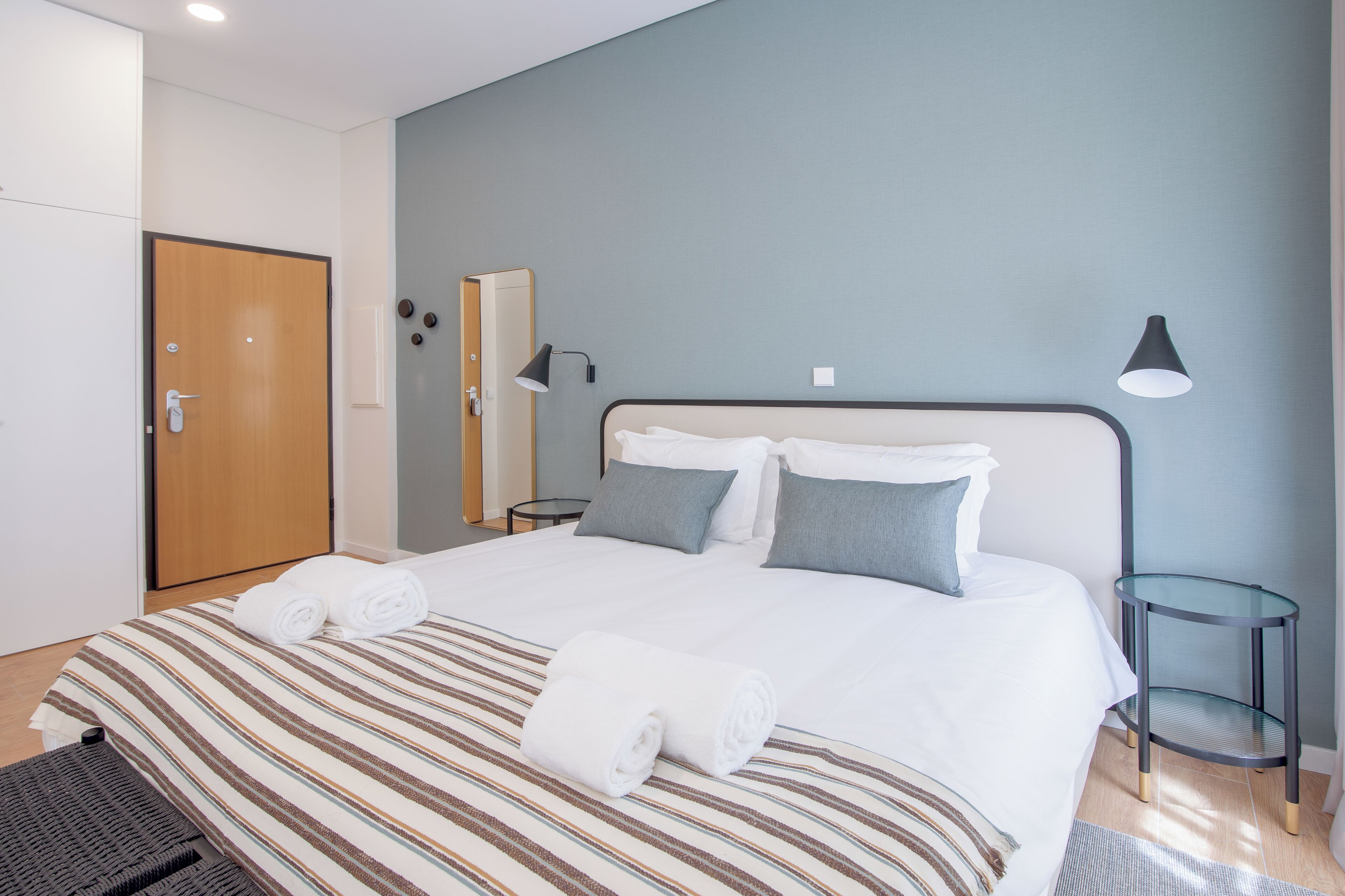 Ein ordentlich bezogenes Bett mit gestreifter Bettwäsche und aufgerollten Handtüchern, eine Nachttischlampe und ein kleiner Glastisch neben dem Bett.