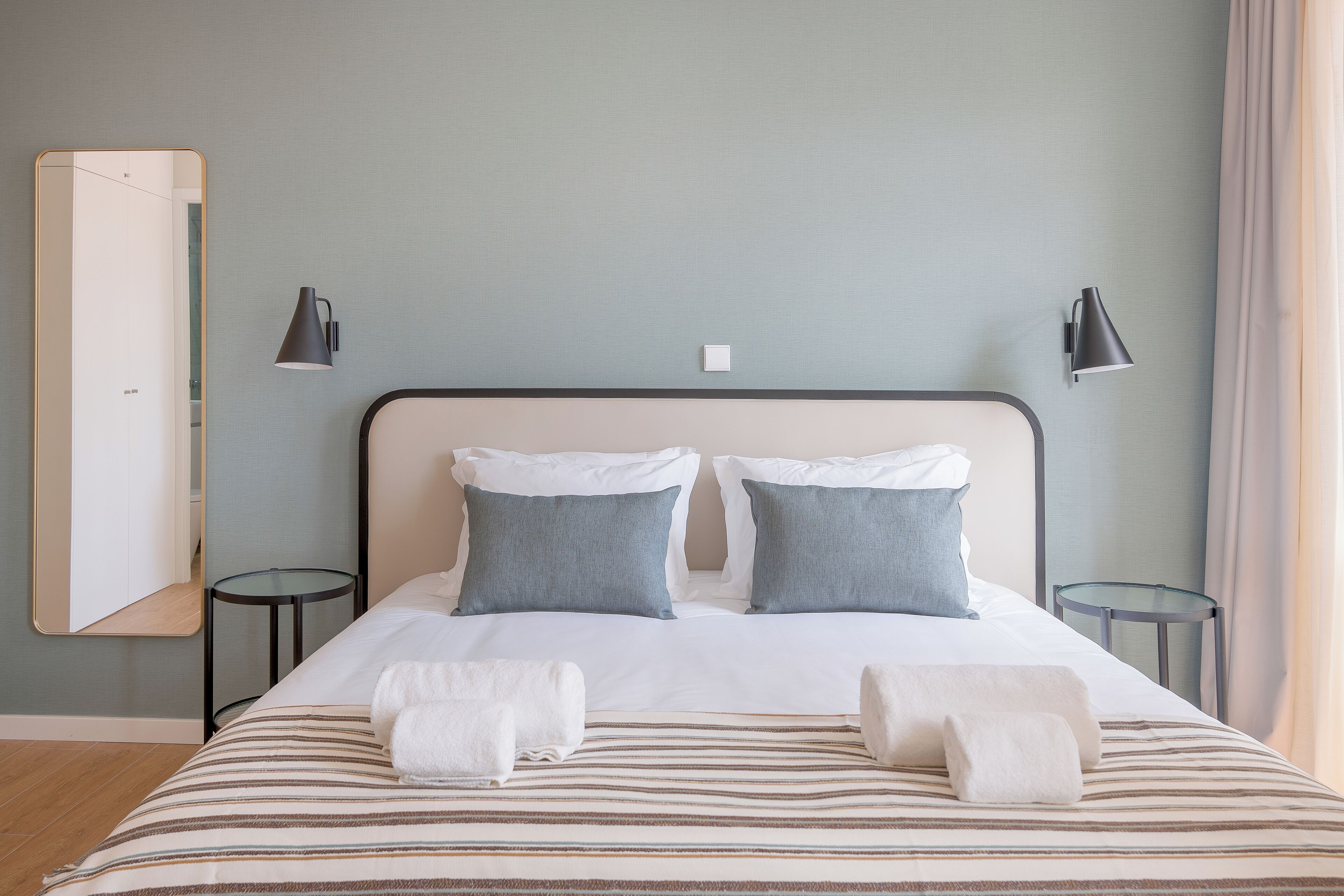 A neatly made bed with striped bedding, two pillows, and two towels. There are two bedside tables, each with a lamp. The room has a light blue wall and a wooden floor.
