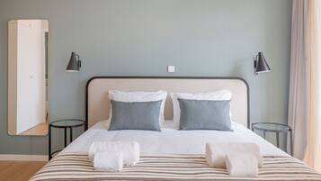 A neatly made bed with striped bedding, two pillows, and two towels. There are two bedside tables, each with a lamp. The room has a light blue wall and a wooden floor.