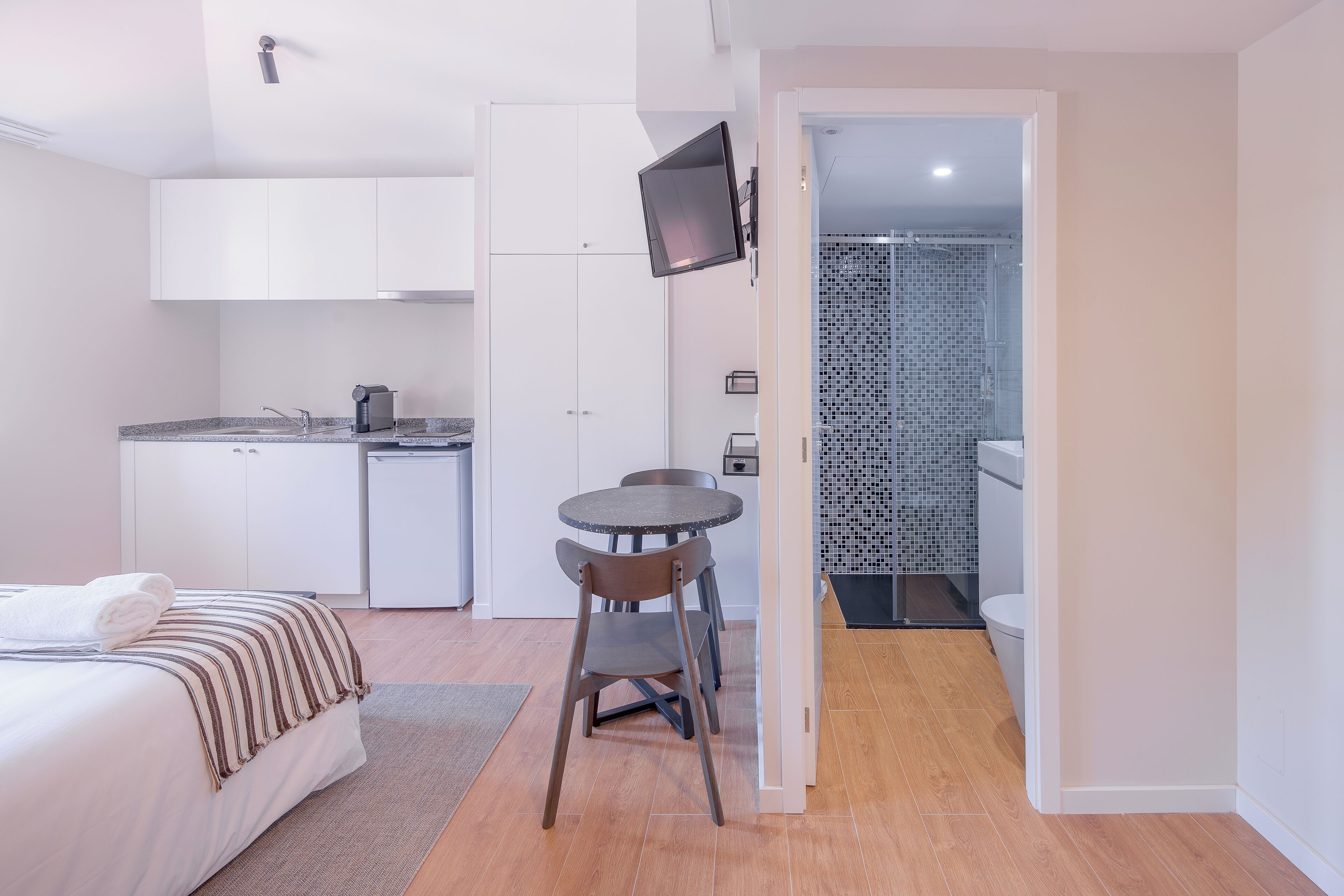 A compact hotel room with a bed, a small dining table with chairs, a kitchenette, and a bathroom visible through an open door.