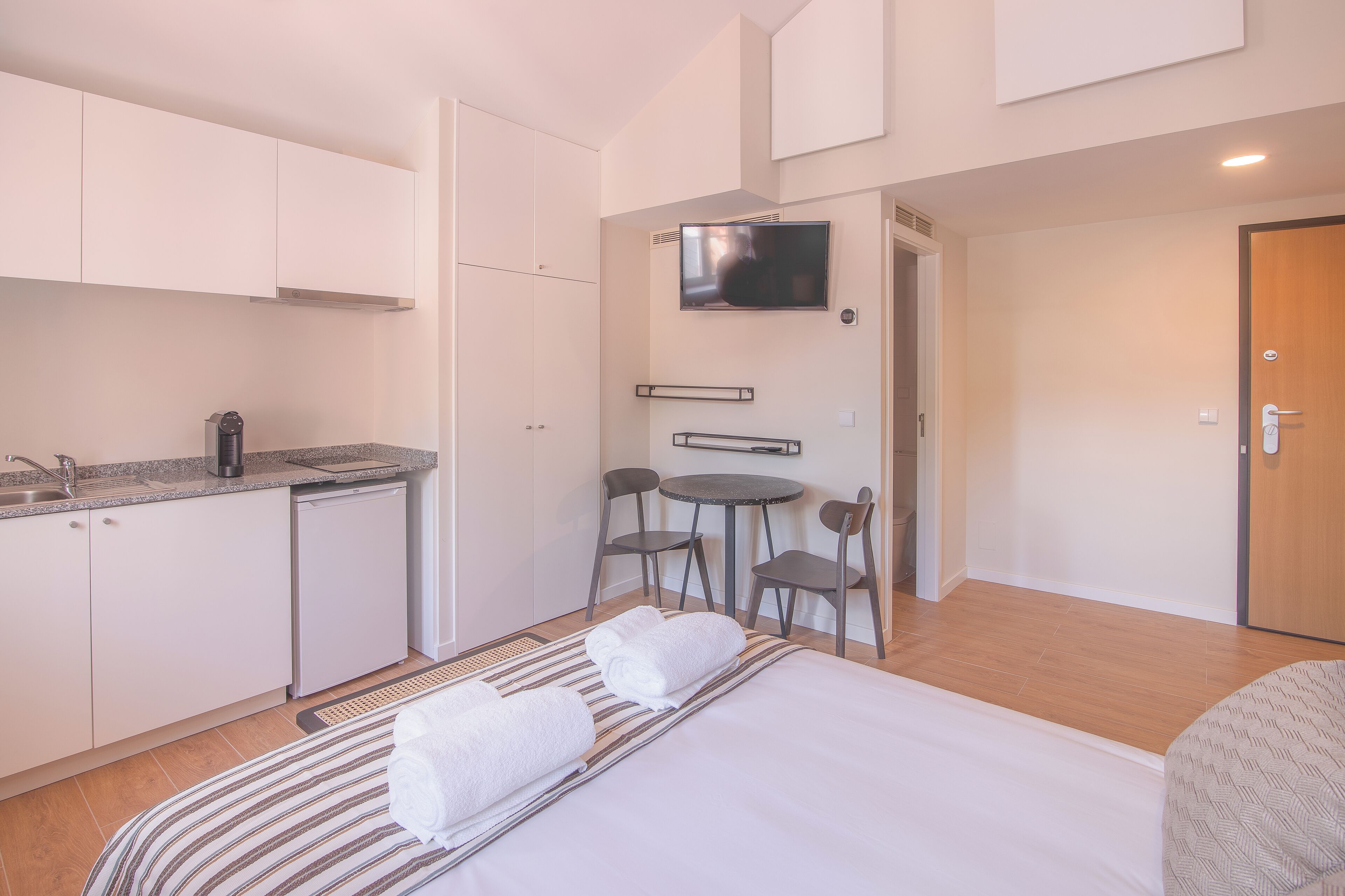 A compact hotel room with a kitchenette, a bed with two rolled towels, a small dining table with chairs, and a TV mounted on the wall.