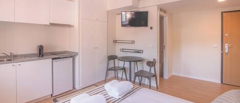A compact hotel room with a kitchenette, a bed with two rolled towels, a small dining table with chairs, and a TV mounted on the wall.