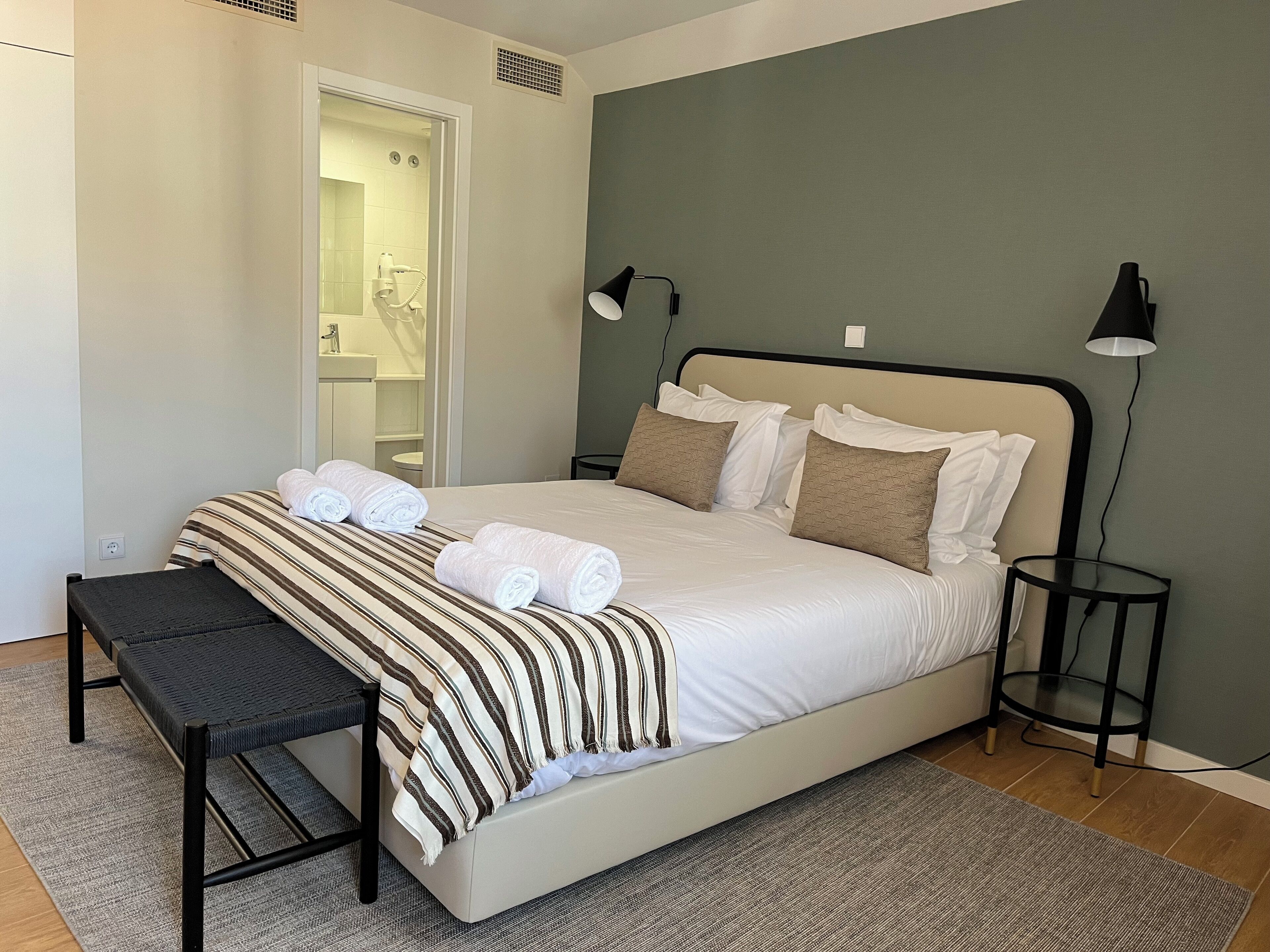 A neatly made bed with white linens and striped blankets, a bedside table, a small round table, and a bench.