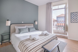A neatly made bed with white linens and striped bedding, a bedside table with a lamp, and a view of a balcony with a railing.