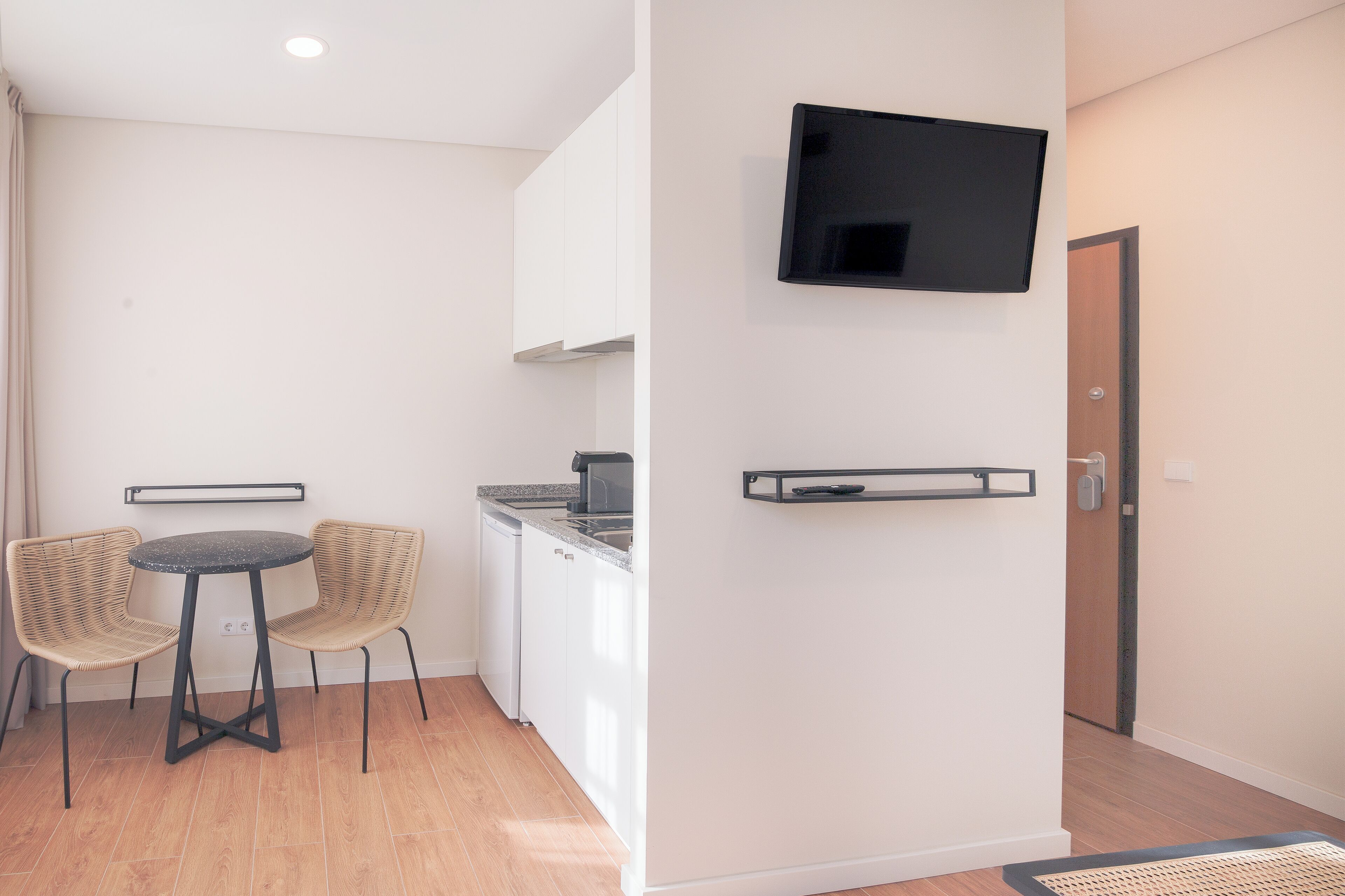 A modern kitchen with a round table, two wicker chairs, a built-in stove, and a flat-screen TV mounted on the wall.