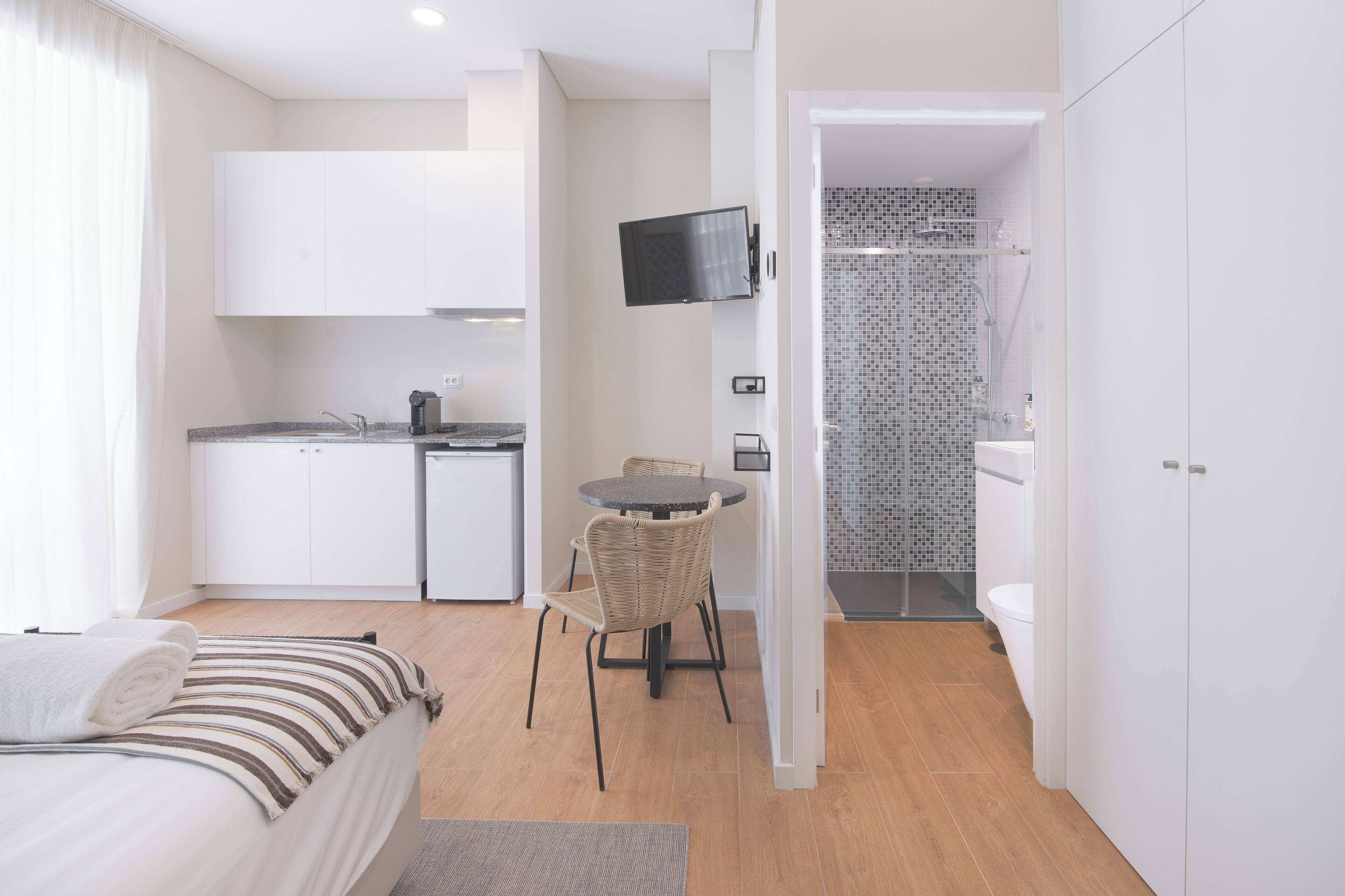 A compact hotel room with a bed, a small kitchenette, a dining area with a table and chairs, a TV mounted on the wall, and a shower area.