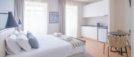 A modern hotel room with a large bed, a small kitchen, a dining area, and a view of the city.