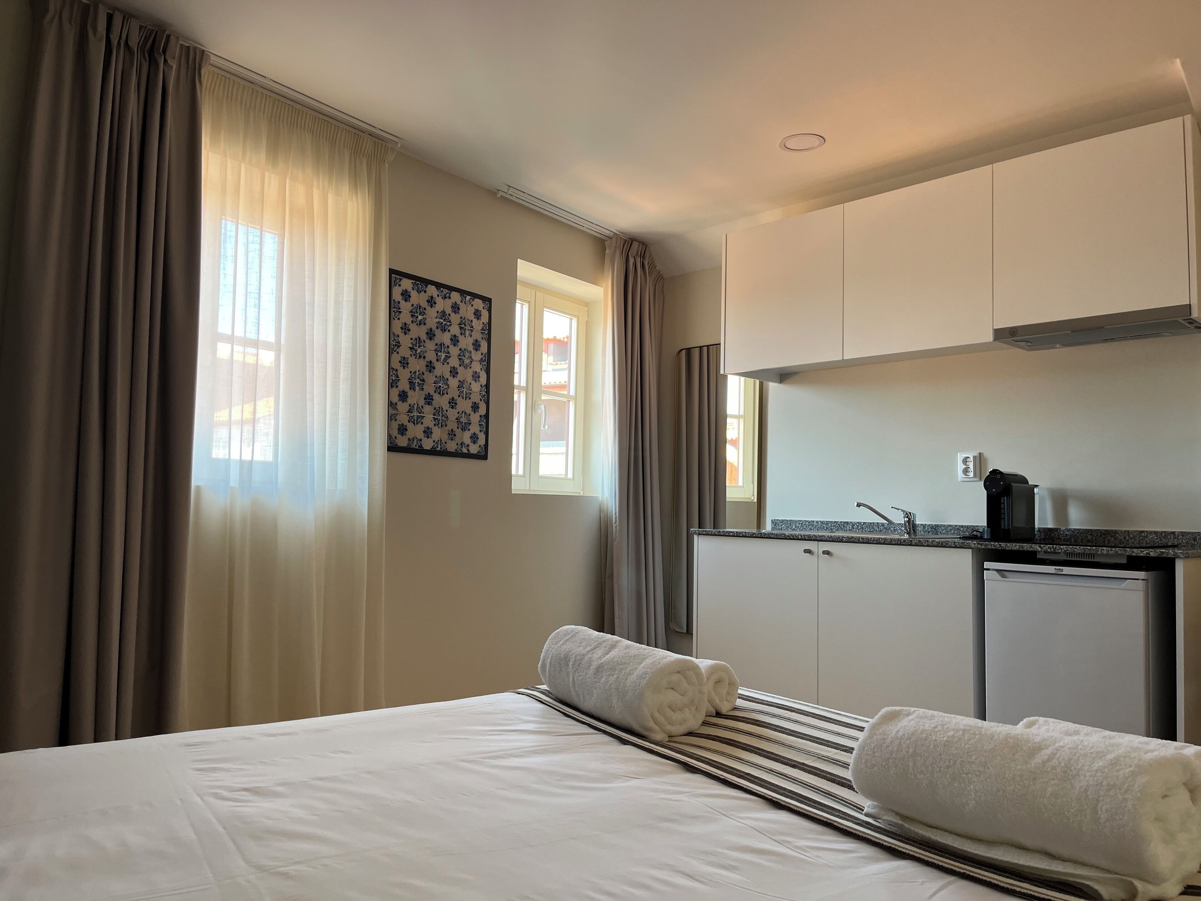 A hotel room with a bed, two rolled towels, a striped blanket, a small kitchenette with a coffee maker, and a window with curtains.