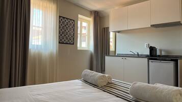 A hotel room with a bed, two rolled towels, a striped blanket, a small kitchenette with a coffee maker, and a window with curtains.