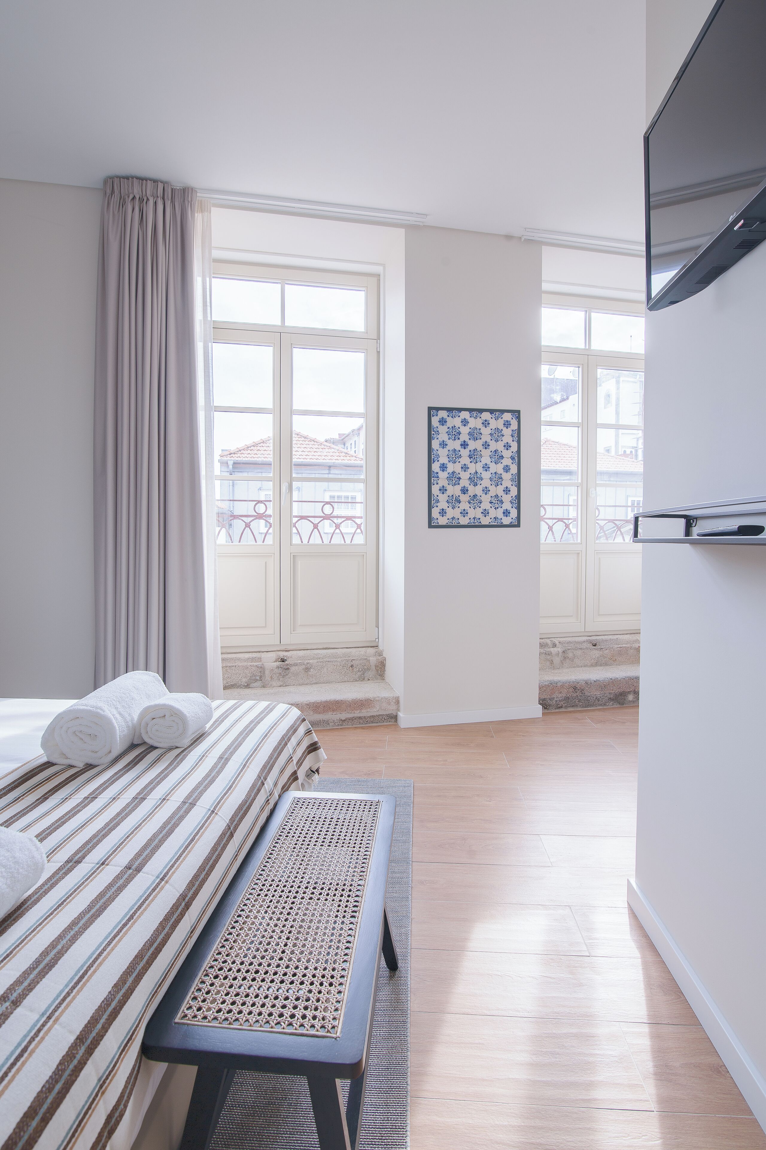 Una habitación de hotel con cama, un banco, televisor, ventana con cortinas y vistas al balcón.