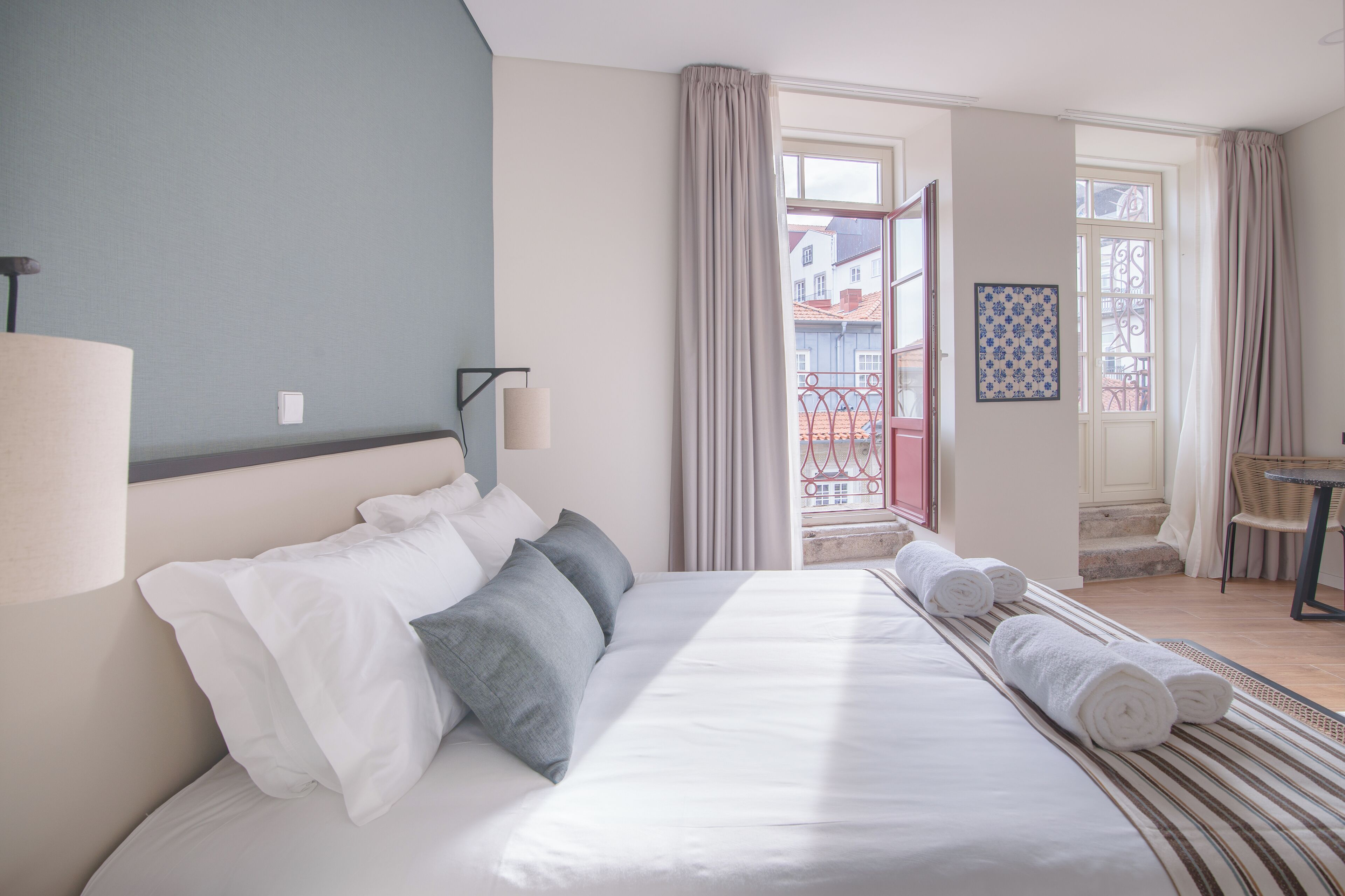 A hotel room with a bed, a lamp, a window with curtains, a chair, and a small table.