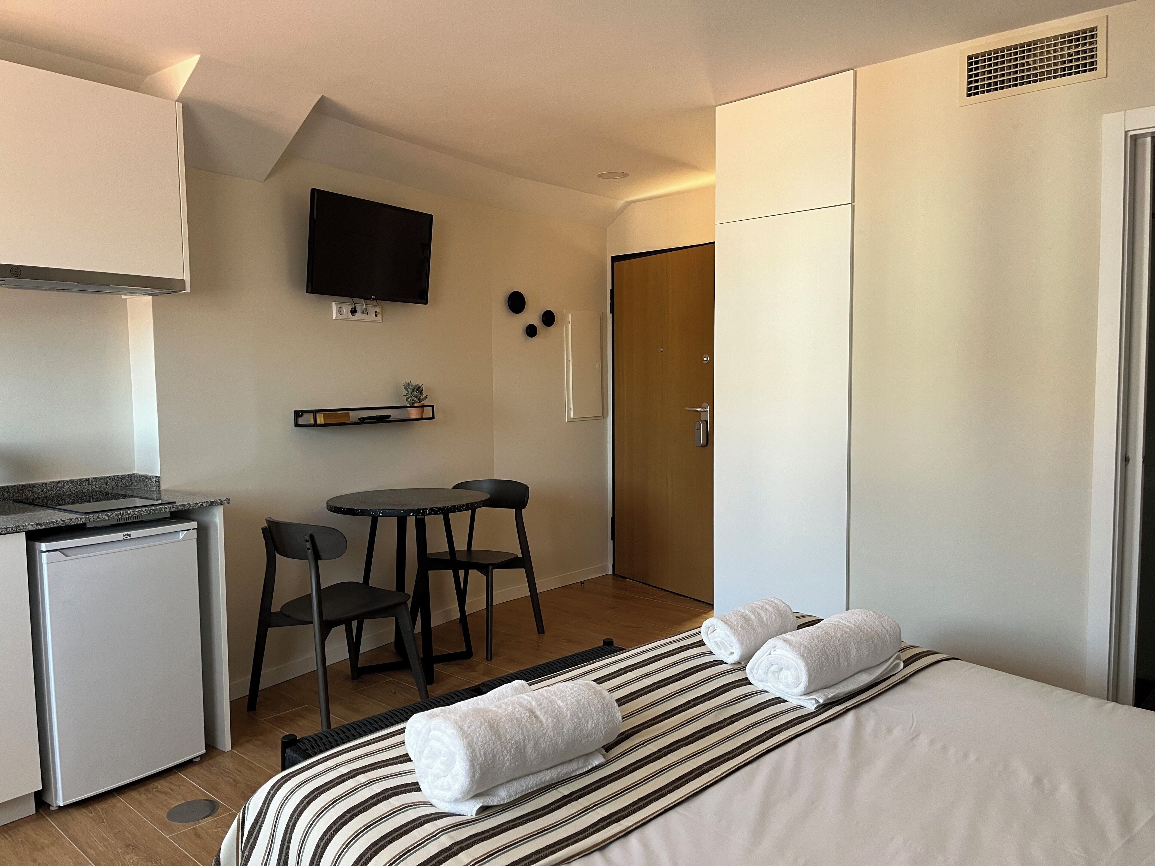 A compact hotel room with a kitchenette, a single bed with two rolled towels, a small dining table with two chairs, a TV mounted on the wall, and a wardrobe.
